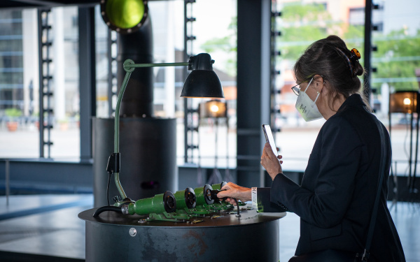 A woman stands in front of an installation. With one hand she inserts the plug belonging to the installation into one of the tube sockets. With the other hand, she photographs the installation with her cell phone.