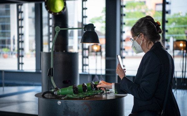 Eine Frau steht vor einer Installation. Mit einer Hand steckt sie den zur Installation gehörenden Stecker in eine der Röhren-Steckdosen. Mit der anderen Hand fotografiert sie ebene jene mit ihrem Handy.