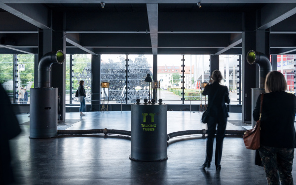 A room whose walls are made of glass. In the center is an installation made of a metal cylinder with the inscription "Talking Tubes". Two tubes connect the cylinder shape to the left and right, each with a large tube.