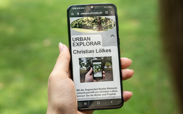 The picture shows a hand holding a smartphone with the work »UrbanExplorAR« on it.
