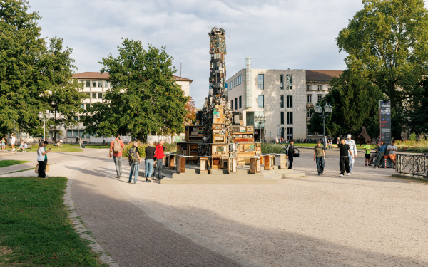 The picture shows the media installation "STREAMERS - a COVID Sculpture" by Benoît Maubrey on Friedrichsplatz. A large tower of old loudspeakers rises into the air and many people have gathered around it to watch it.