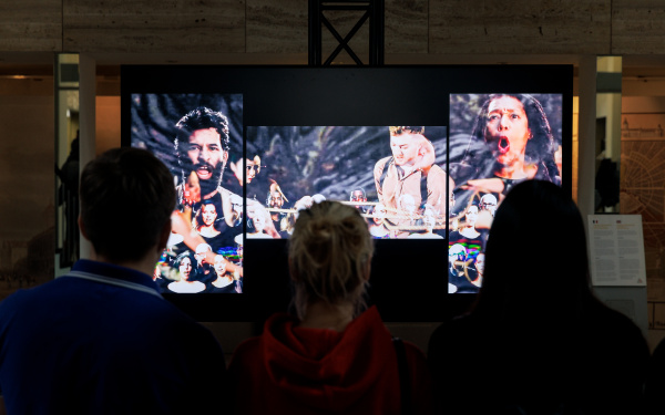 Auf dem Bild ist die Videoinstallation von John Sanborn im Foyer des rathauses zu sehen. Aud 3 Bildschirmen sind mehrere Menschen abgebildet, die unterschiedliche Expressionen ausdrücken.