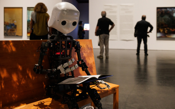 Zu sehen ist ein Roboter, dessen Aufbau dem Korpus des Menschen gleicht. Er sitzt auf einer Bank.