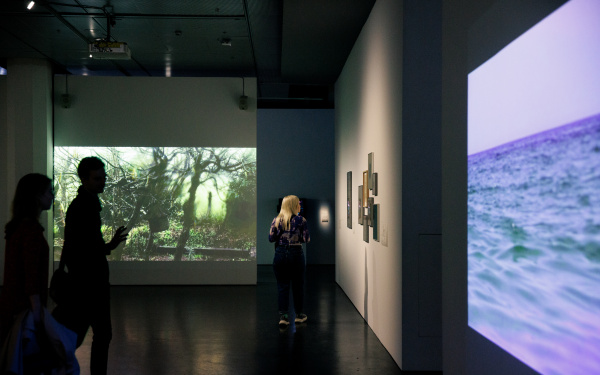 Zu sehen ist die Ausstellung »Soun-Gui Kim: Lazy Clouds« am ZKM. Hinten sind Aufnahmen der Natur zu sehen, rechts sieht man Aufnahmen eines Meers.