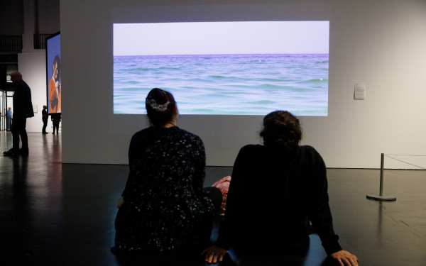 Zu sehen sind zwei sitzende Personen vor einem Bildschirm. Auf dem Bild ist das Meer zu sehen.