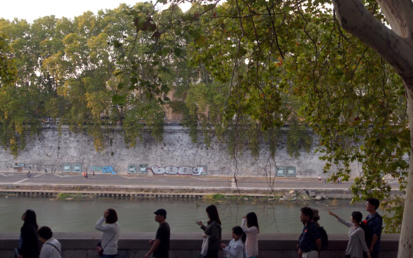Film still by Marijke van Warmerdam. You see a riverbank in Rome. On the right a tree and on the left people walking along the riverbank.
