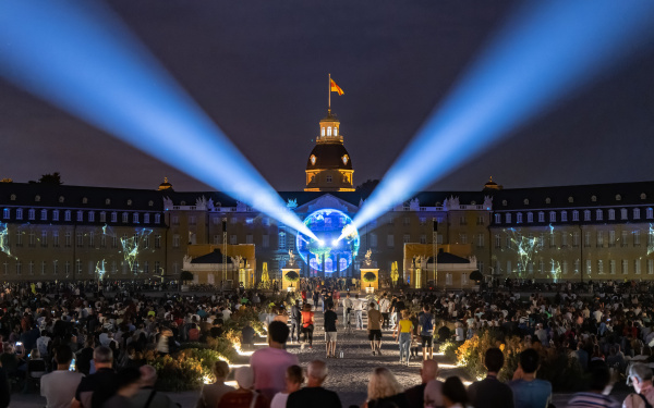 Auf der Fassade des Karlsruher Schlosses leuchtet die Erdkugel auf. Zwei Strahler leuchten über das Publikum hinweg.