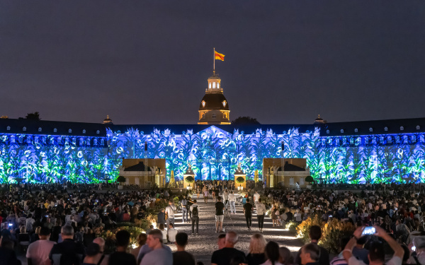 Das Karlsruher Schloss erleuchtet in einem blau-grünen Pflanzenmuster. Zu sehen ist auch die vielen Menschen, die im Publikum sitzen.