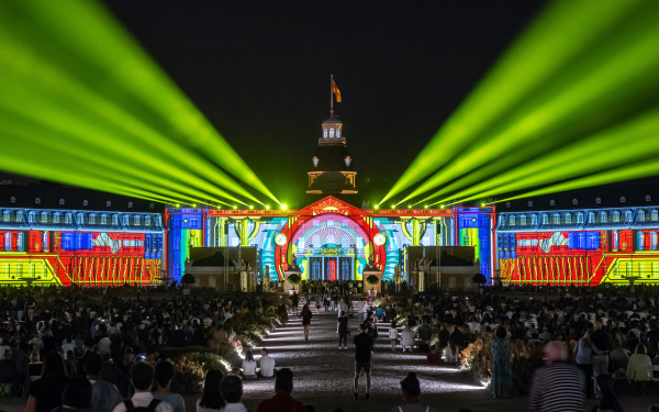 In diesem Bild erleuchtet das Karlsruher Schloss kunterbunt in einem Comic-Stil. Gelbe Strahler leuchten in den Schwarzen Himmel. Die Zuschauer füllen den rasen vor dem Schloss aus.