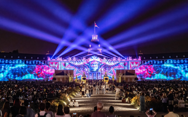 The castle lights up in a color combination of blue, purple and pink tones. Depicted is a face under a mask, which runs outward in geometric shapes. Purple spotlights illuminate the sky.