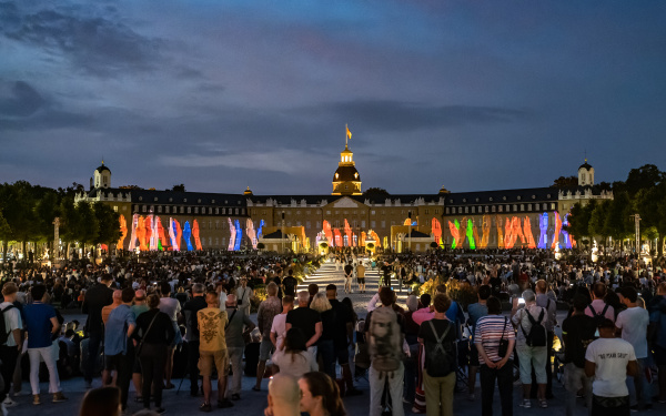 Im Vordergrund sieht man das Publikum das bei der Eröffnung der Schlosslichtspiele anwesend war. Auf dem Schloss projeziert sieht man lauter bunte Hände, die vom Boden nach oben ragen.