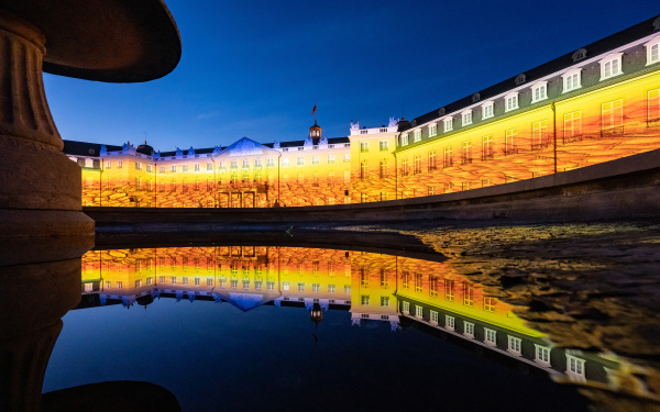 The castle of Karlsruhe illuminates in yellow-orange hues. In the foreground the facade is reflected in the water of the fountain. The evening sky has already darkened.