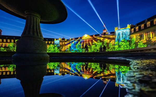 In the background you can see the illuminated facade of Karlsruhe Castle. A blue sky is projected over a landscape of trees with a waterfall. In the foreground the projection is reflected in a fountain. Spotlights rise into the sky.