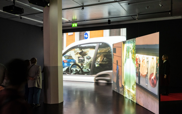Ausstellungsansicht »Marijke van Warmerdam. Then, now, and then«. Zu sehen sind zwei Personen vor zwei großen Bildschirmen. Auf einem davon ist eine Nahaufnahme von einem Auto zu sehen.