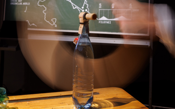 In the foreground there is a glass bottle on which a kind of tube was fixed. In the background there is a board with statistics.