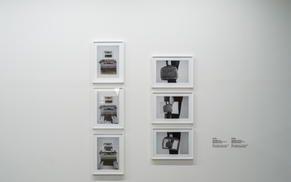 Zu sehen sind sechs Bild-in-Bild, schwarz-weiß Fotografien in Bilderrahmen an einer Wand. Drei von ihnen zeigen eine Schreibmaschine, die anderen drei zeigen eine Hand, die eine Aktentasche hält.