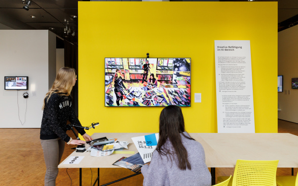 Zu sehen sind zwei Personen, eine stehend, eine sitzend, an einem Tisch. An der Wand vor ihnen hängt ein Bildschirm mit einer Kamera. Auf dem Bildschirm ist in abstrakter und künstlerischer abgebildet, was die Kamera aufzeichnet.