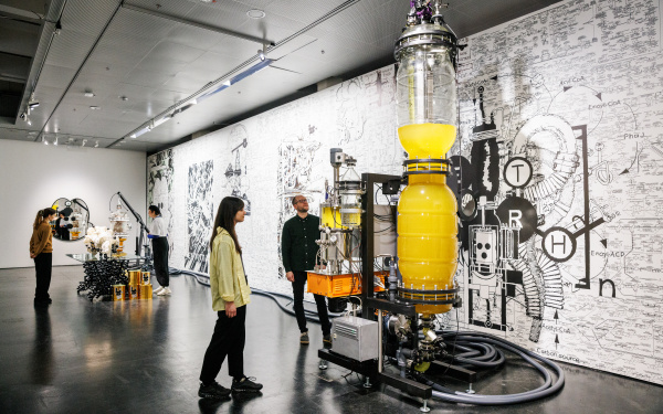 A ceiling-high, elongated glass vessel containing a yellow liquid can be seen. Several cables or hoses lead to smaller glass vessels that also contain liquid. Large hoses are lying on the floor. Two people are looking at this.