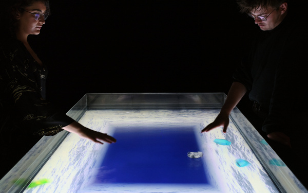 On display is the work "A volve". The picture shows two people looking at the work from one side each. This is on a screen that lies horizontally and can be controlled with touches.
