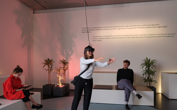 Zu sehen ist das Werk »ARtchive«. Das Bild zeigt drei Personen, zwei sitzen auf den Sofas und eine Person steht in der Mitte und hat eine VR-Brille auf und Interagiert mit etwas, das nur Sie sehen kann.