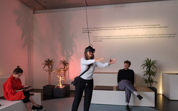 On display is the work "ARtchive". The picture shows three people, two sitting on the sofas and one person standing in the middle with VR glasses on and interacting with something that only she can see.