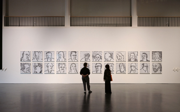 Das Bild zeigt eine Totalaufnahme des Werkes »Porträt on the Fly«, vor dem zwei Personen mit dem Rücken zur Kamera stehen und die Bilder betrachten. Im gesamten sind zwei Reihen mit je 12 Bildern zu sehen, die alle das Porträt einer Person zeigen.