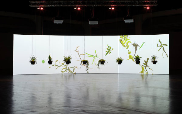 The frontal view of the artwork "Eau de Jardin" is shown. In the foreground, some plants hang in a semicircle from the ceiling and in the background a large canvas can be seen on which there are floral shapes. 