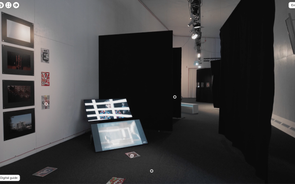 View into an exhibition room, which is hung with room-dividing black curtains. On the left side of one wall hang small photographs of paintings and colorful drawings with large letters. Two screens in the room show various videos, apparently of performanc