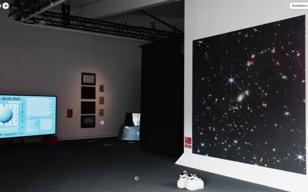 View of an exhibition room, with a large screen with informational text on the left, and several paintings and photographs on the wall next to it. On the right side of the exhibition space is a large image with twinkling stars in black space. On the floor