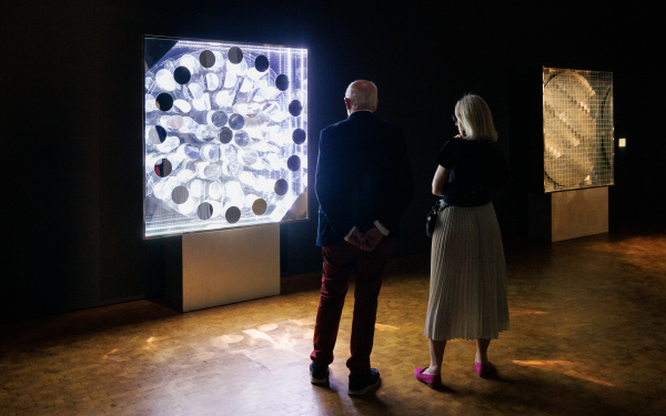 Zu sehen sind zwei Menschen, die vor einem Leuchtkasten stehen, der an der Wand hängt. Durch verschiedene Objekte in den Kasten bricht das Licht und es entstehen optische Variationen.