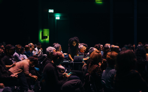 Jessica Ekomane während einer Performance an ihrem Laptop stehend zwischen einer sitzenden Menschenmenge