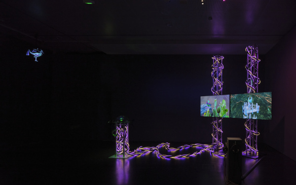 Auf dem Bild ist ein dunkler Raum zu sehen. Rechts im Bild befindet sich ein Kunstwerk/ Installation, bestehend aus zwei Bildschirmen, die von Leuchtelementen umgeben sind.