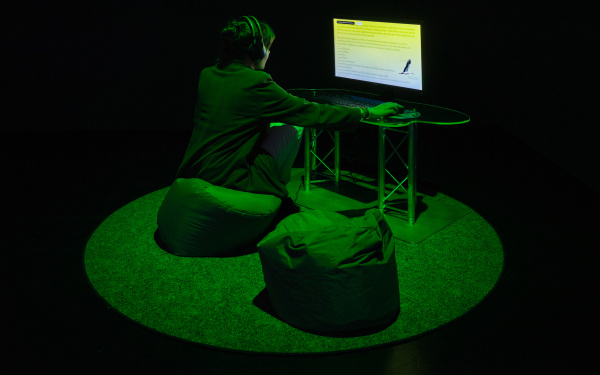 The picture shows a dark room with a green illuminated area in the middle. In this area there is a computer in front of which a young woman is sitting in a beanbag and looking at the computer screen.