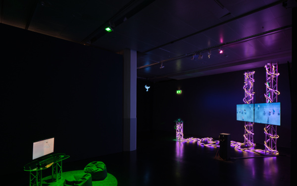 The picture shows a dark room with two works of art in the middle. On the left is a green-lit space with a computer and two beanbags and on the right is an installation with two screens surrounded by light elements. 