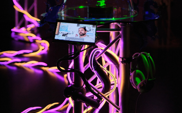 In a close-up, a kind of cylinder can be seen, illuminated by strands of pink-pink. In the background, the rest of the artwork is blurred, the focus is on the glass cylinder to which a small screen is attached on which a man seems to be speaking.