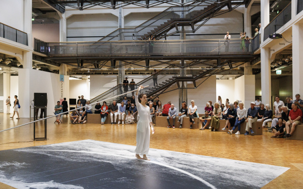 Performance von Ulrike Rosenbachs »Die einsame Spaziergängerin«, dir Künstlerin, ganz in weiß gekleidet hält eine Stange fest und läuft im Halbkreis einen Weg ab, während sie gefilmt wird. Im Hintergrund sieht man ein großes Publikum.