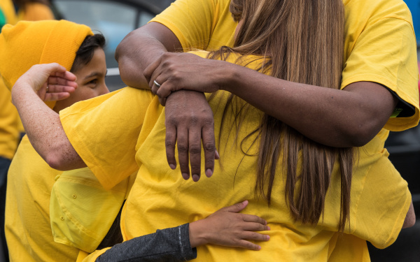 Eine Frau in einem gelben T-Shirt umarmt ein Kind, welches ebenfalls gelb trägt, die Gesichter sind nicht zu sehen. Im Fokus stehen die Arme, die sich umschlingen.