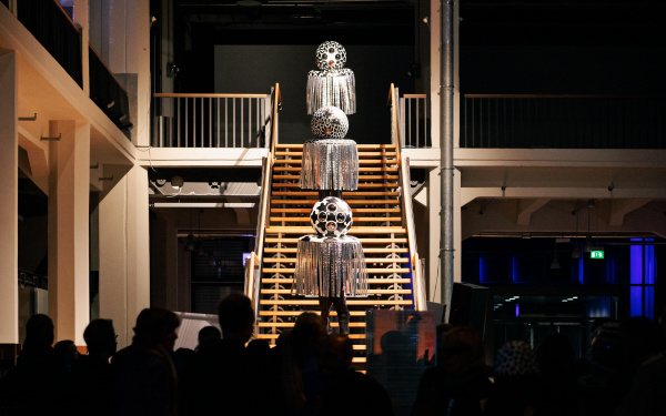 Auf der Treppe in dem Foyer des ZKM läuft eine Performance. Im Abstand von einigen Stufen stehen drei menschen auf der Treppe und tragen eine Kugel über ihrem Kopf, an der Schnüre hinuterhängen.