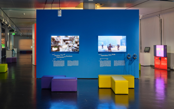 Blaue, freistehende Wand in der Ausstellung »Playful Architectures«. Daran angebracht sind zwei Bildschirme, die Computerspiel zeigen. Im restlichen Ausstellungsraum verteilen sich weitere Bildschirme.