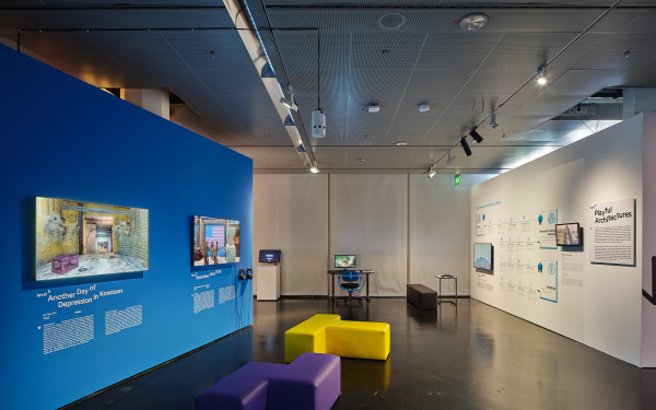 Blaue Wand in der Ausstellung »Playful Architectures« auf der linken Seite. Daran angebracht sind zwei Bildschirme, die Computerspiel zeigen. Im restlichen Ausstellungsraum verteilen sich weitere Bildschirme.