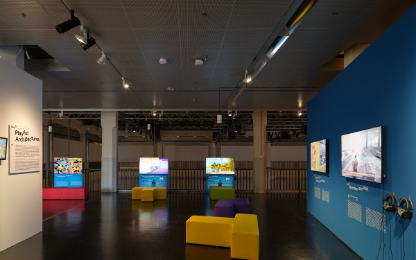 Blaue Wand in der Ausstellung »Playful Architectures« auf der rechten Seite. Daran angebracht sind zwei Bildschirme, die Computerspiel zeigen. Im hinteren Ausstellungsbereich verorten sich weitere Bildschirme. Links steht eine weitere Ausstellungswand.