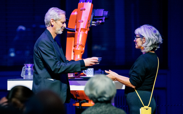 Im Hintergrund ist ein orangener Roboter, der gerade tee gemacht hat, den der künstlerisch-wissenschaftliche Leiter des ZKM | Karlsruhe Alistair Hudson einer Frau überreicht.