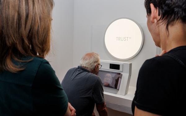 Drei Besucher:innen beschäftigen sich mit der interaktiven Installation "TRUST". Zwei Besucherinnen beobachten, wie ein Besucher vor dem Avatar sitzt. 