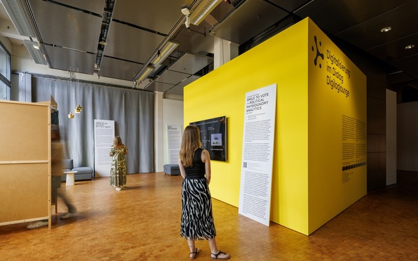 Ausstellungsansicht mit drei Personen im Raum, die Wandtexte lesen und sich mit einer interaktiven Installation beschäftigen