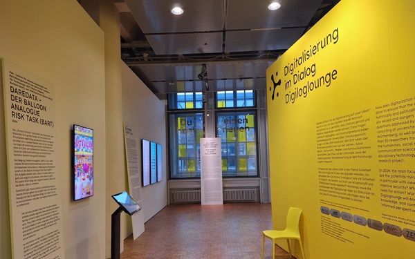 Das Bid zeigt den Eingang in die Digiloglounge. Rechts ist eine beschriftete gelbe Wand, links steht eine interaktive Installation bestehend aus einer freistehenden Touchtastatur und einem Wandmonitor. Im Hintergrund sind die mit dem Ausstellungstitel beschrifteten Fenster zu sehen.