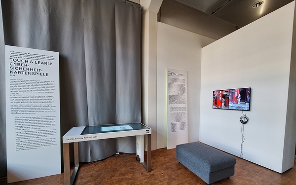 Ausstellungsansicht. Zu sehen sind ein interaktiver Touchtable auf dem digitale Kartenspiele gespielt werden können und ein Wandmonitor auf dem eine Videoarbeitabspielt. 
