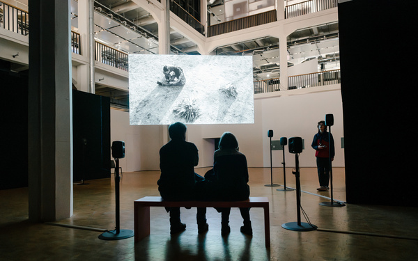 Two people are seated on a bench in front of a screen. 