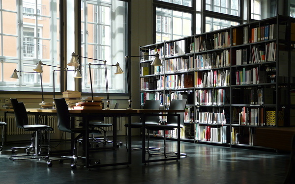 Zu sehen ist ein Tisch mit Stühlen sowie ein Bücherregal. 