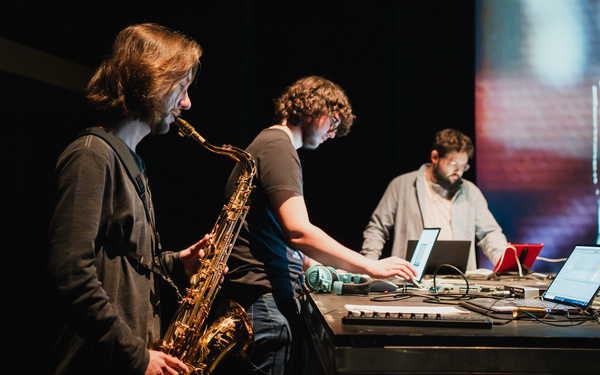 Drei Musiker stehen auf einer Bühne, einer von ihnen spielt Saxophon.
