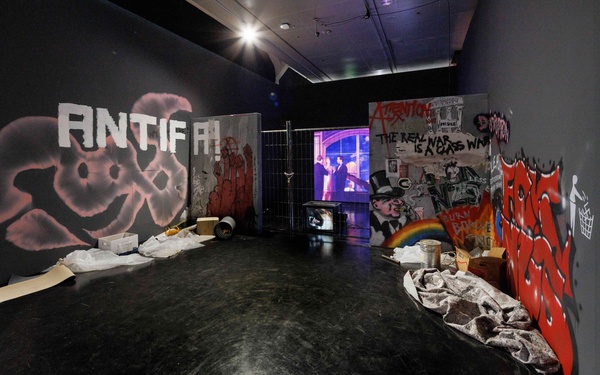 Installation in einem Ausstellungsraum mit politischen Graffiti, Absperrgitter, Projektionsbildschirm und verstreuten Materialien auf dem Boden.